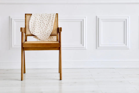 Ein Holzstuhl mit einer geflochtenen Rückenlehne und Sitzfläche steht auf einem weißen Boden vor einer weiß getäfelten Wand. Ein cremefarbener Stoff mit kleinem Blumenmuster ist über den Stuhl drapiert.