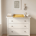 In einem minimalistischen Kinderzimmer steht eine weiße Kommode mit drei Schubladen und dem KraftKids Wickelaufsatz weiß für 108 cm breite HEMNES Kommode (Wickelfläche 50 x 70 cm), darüber eine gelbe Wickelauflage, gefaltete Handtücher und eine Flasche. Darüber hängt begrünte Kunst, durch die das Sonnenlicht fällt.