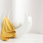 Ein moderner weißer halbmondförmiger Schaukelstuhl mit einem runden strukturierten Kissen hat die KraftKids Babydecke Leinen gelb senfgelb über eine Seite drapiert. Der Hintergrund besteht aus weißen vertikalen Paneelen und einem hellen Holzboden.