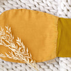 Ein handschuhförmiger KraftKids Strampelsack Frühling Sommer aus Leinen in senfgelb liegt auf grob gestricktem, weißem Untergrund und ist mit zwei Trockenblumen dekoriert.