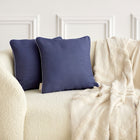 Ein cremefarbenes Struktursofa mit zwei KraftKids Kissenbezug Leinen in Dunkelblau und einer hellbeigen Überwurfdecke über der Armlehne, vor einer weiß getäfelten Wand.