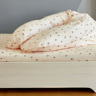 Ein ordentlich gemachtes Bett mit hellen Laken und einem KraftKids qualitäts Stillkissen Musselin Kirschen steht vor einer grauen Wand und einem Holzboden, ergänzt durch einen ergonomisch hellen Holzbettrahmen für eine gemütliche Szene.