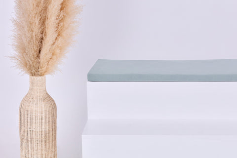 Eine hohe geflochtene Vase mit beigem Pampasgras steht neben einer minimalistischen weißen Bank, die mit dem Kissen KraftKids GmbH Sitzauflage für Ikea STUVA/SMÅSTAD Musselin mint vor einem schlichten hellen Hintergrund belegt ist.