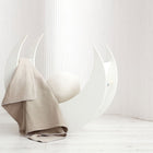 Eine moderne weiße Mondsichelwiege mit einem cremefarbenen Kissen ist in der KraftKids Babydecke Musselin beige drapiert und steht vor einem minimalistischen Hintergrund mit weißem Holzboden und dezent gestreiften Wänden.