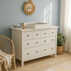 Eine weiße HEMNES-Kommode mit dem KraftKids Wickelaufsatz weiß zum Quer-Wickeln steht vor einer blauen Kinderzimmerwand, die von einem Löwengesicht gekrönt wird. In der Nähe stehen ein Korbstuhl, eine Topfpflanze, Holzspielzeug und ein runder Teppich.