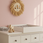 Ein Kinderzimmer mit einer rosafarbenen Wand, einem Wandbehang mit Löwengesicht und einem KraftKids Wickelaufsatz weiß zum Quer-Wickeln auf einer weißen HEMNES Kommode mit dunklen Knöpfen. Ein Holzspielzeugpferd und gefaltete Tücher liegen daneben. Sonnenlicht wirft Rechteckschatten an die Wand.