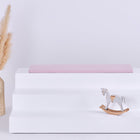 Eine minimalistische Szene zeigt ein weißes dreistufiges Podest mit einem KraftKids GmbH Sitzauflage für Ikea TROFAST Doppelkrepp Rosa Kissen, ein hölzernes Schaukelpferd davor und eine hohe geflochtene Vase mit Pampasgras auf der linken Seite vor einem hellen Hintergrund.
