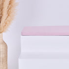 Eine hohe geflochtene Vase mit beigem Pampasgras steht neben einer modernen weißen Bank mit der KraftKids GmbH Sitzauflage für Ikea TROFAST Doppelkrepp Rosa vor einem schlichten weißen Hintergrund.