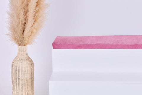 Eine hohe geflochtene Vase mit beigem Pampasgras steht neben einem weißen Stufenpodest, auf dem das Kissen Cord Breitcord Rosa der KraftKids GmbH Sitzauflage für Ikea STUVA/SMÅSTAD liegt. Der schlichte, helle Hintergrund unterstreicht die minimalistische, heitere Stimmung.