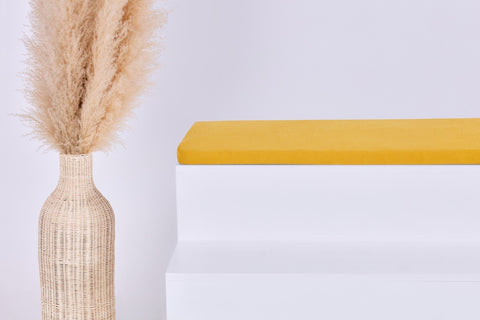 Eine hohe geflochtene Vase mit beigem Pampasgras steht neben einem modernen weißen Stufenpodest, das mit dem KraftKids GmbH Sitzauflage für Ikea STUVA/SMÅSTAD Doppelkrepp Gelb Mustard Kissen belegt ist. Der Hintergrund ist schlicht und hell gehalten.