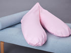 Das KraftKids Stillkissen "kleine Blätter rosa auf Weiß" mit ergonomischer Form liegt auf einer hellblauen Polsterbank mit Holzbeinen vor grauem Hintergrund.