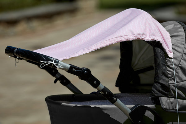 Nahaufnahme eines grauen Kinderwagens mit dem KraftKids Sonnensegel kleine Blätter rosa auf Weiß, das mit weißen Schnüren an den Griffen befestigt ist und den Kinderwagen an einem sonnigen Tag vor einem unscharfen Hintergrund im Freien schützt.