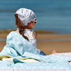 Ein junges Mädchen mit Kopftuch und Sonnenbrille sitzt auf der KraftKids Babydecke weiße Diamante auf Pastell Blau am Strand und blickt mit dem Rücken zur Kamera aufs Meer. Der Sandstrand und das ruhige blaue Wasser sind im Hintergrund zu sehen.