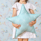 Ein junges Mädchen in einem weißen Ösenkleid hält das KraftKids Sternkissen "weiße dünne Diamante auf Mint" in der Hand und steht vor einer pastellfarbenen, vogelgemusterten Kinderzimmerwand, das Gesicht verborgen.