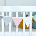 Ein weißes Kinderbett wird mit der KraftKids Wimpelkette weiße dünne Diamante in Pastellgrau, Rosa, Weiß und Teal geschmückt, wobei jede Flagge geometrische Muster aufweist. Gemütliche Kissen vervollständigen diese charmante Kinderzimmerdekoration.