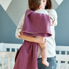 Eine Frau hält ein Baby, das in eine KraftKids Babydecke Musselin purpur eingewickelt ist, und steht neben einem weißen Kinderbett in einem modernen Raum mit geometrischen Wandmustern.