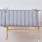 Ein graues Kinderbett mit dem KraftKids Bettumrandung Nestchen Musselin grau Pusteblumen aus weichem Musselin steht auf einem hellen Holzgestell vor einer weißen Wand mit Zierleisten.