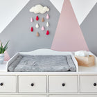 Ein weißer KraftKids Wickelaufsatz XXL extra breit mit Trennfach für HEMNES mit einer grau gemusterten Wickelauflage, einer Topfpflanze und einer Windelbox aus Holz. An der Wand befinden sich rosa und graue geometrische Formen und ein Wolkenmobile mit Regentropfen.