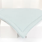 Ein grüner, rautenförmiger KraftKids Wickelauflage-Musselin mit kleinen goldenen Punkten wird auf einer weißen, geometrischen Fläche in einem minimalistischen Rahmen präsentiert.