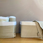 Zwei KraftKids Körbchen Leinen Natur Braun stehen auf einer Holzfläche, ideal für die Aufbewahrung am Wickeltisch. In dem einen Körbchen liegen gerollte weiße Handtücher, über den Rand des anderen ist ein gefaltetes Tuch drapiert. Von rechts scheint sanftes Licht herein.