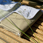 Zwei fein säuberlich gefaltete beige-weiße Handtücher liegen auf einer hölzernen Lattenbank. Dünnes grünes Gras wirft im hellen Sonnenlicht Schatten auf die KraftKids Wickelunterlage Leinen Natur Braun.