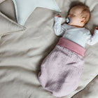 Ein Baby schläft friedlich in einem Bett, trägt ein weißes Oberteil und den KraftKids Strampelsack Herbst Winter Musselin in goldene Punkte auf Rosa, mit einem Schnuller. Weiche sternförmige Kissen und neutrales Bettzeug umgeben das Baby.