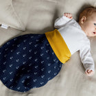 Ein Baby liegt auf einem beigen Bett in einem KraftKids Strampelsack Herbst Winter Musselin dunkelblau Anker, mit weißen Ankern. Das mit einem weißen Oberteil bekleidete Baby blickt zur Seite, ein Arm angewinkelt, der andere ruht an der Seite.