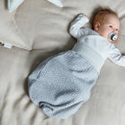 Ein Baby, das den KraftKids Strampelsack Herbst Winter Musselin grau Punkte trägt, liegt auf einem hellen Bett mit einem Schnuller und schaut zur Seite. Der kuschelige graue gepunktete Schlafsack ist perfekt für die kalte Jahreszeit, mit einem Kissen in Sichtweite.