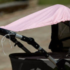 Eine Nahaufnahme eines KraftKids Sonnensegels "weiße Punkte auf Koralrosa" ist mit weißen Schnüren an einem Kinderwagengriff befestigt. Der Baumwollstoff hebt sich ab, während der unscharfe Hintergrund auf den Bürgersteig und das Grün im Freien hindeutet.