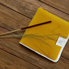 Ein geschlossenes gelbes Notizbuch mit einem weißen Etikett liegt auf einer Holzfläche. Diagonal darüber liegen ein brauner Stift, ein getrockneter Grashalm und die KraftKids Reisewickelunterlage Doppelkrepp Gelb Senf.
