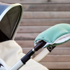 Eine Nahaufnahme des KraftKids Kinderwagenmuffs Doppelkrepp Grün Jade, handgenäht in Grün und Weiß, befestigt an einem Kinderwagengriff. Sein wärmeisolierendes Design hebt sich von dem Hintergrund aus Holzplanken ab.