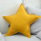 Ein KraftKids Sternkissen Doppelkrepp Gelb Senf in leuchtendem Gelb ruht auf hellgrauen Kissen auf einer hellen Couch mit einer Betonwand - perfekt, um jedem Kinderzimmer einen verspielten Stil zu verleihen.