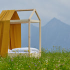 Ein minimalistisches Holzbettgestell mit dem KraftKids Bettbaldachin Musselin gelb Pusteblumen und weißem Bettzeug steht in einem Wildblumenfeld, im Hintergrund erheben sich neblige Berge unter einem wolkenverhangenen Himmel.