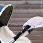 Nahaufnahme eines Kinderwagens mit dem KraftKids Kinderwagenmuff Doppelkrepp Rosa, der für zusätzliche Wärme am Lenker befestigt ist, vor Holzlatten. Eine cremefarbene Decke ist im Inneren des Kinderwagens drapiert.