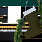 Eine KraftKids Weihnachtssocke Doppelkrepp Grün Herbstgrün hängt an einer mit Tannengirlanden geschmückten Leiter, während im Hintergrund ein verschwommenes Klavier und offene Notenblätter die gemütliche Atmosphäre der Weihnachtsdeko unterstreichen.