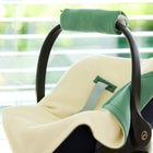 Eine KraftKids Decke für Babyschale Winter Doppelkrepp Grün Jade wird auf einem leeren Kinderautositz in der Nähe eines Fensters mit horizontalen Jalousien gezeigt, wobei das Sonnenlicht einfällt und die zarten Creme- und Grüntöne des Stoffes sichtbar sind.